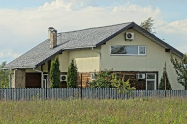 Kiremit çatılı bir evin altında yeşil çimenli gri bir çitin arkasında gökyüzü ve bulutların arkasında kahverengi bir ev.