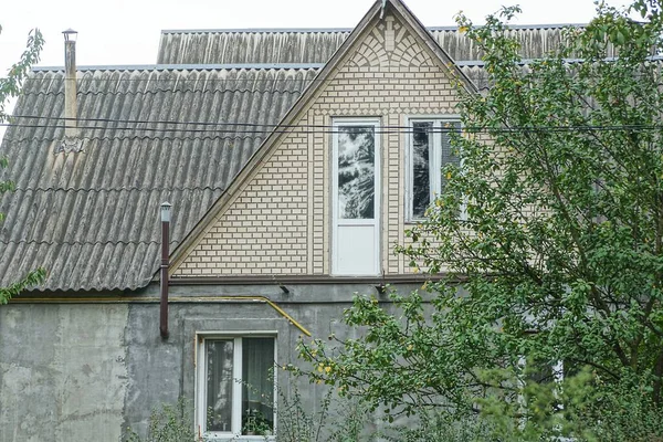 Kırsal kesimdeki gri beton bir evin ön cephesinde kahverengi tuğlalı bir tavan penceresi ve yeşil bir ağacın arkasındaki çatı altında bir kapı var.