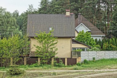 Çitin arkasındaki yeşil bitki örtüsünün altında penceresi ve bacası olan kahverengi bir özel ev.