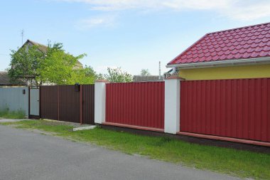 Kahverengi metal bir kapı, kırmızı demir bir çit duvarında. Sokakta yeşil çimlerde beyaz beton sütunlar var.