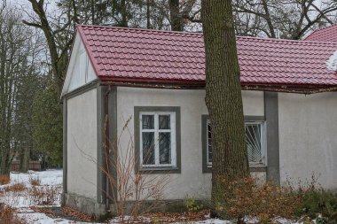 Kırmızı kiremitli çatısı altında pencereleri olan gri özel bir evin ön cephesi beyaz karlı bir kış sokağında ağaçların arasında.