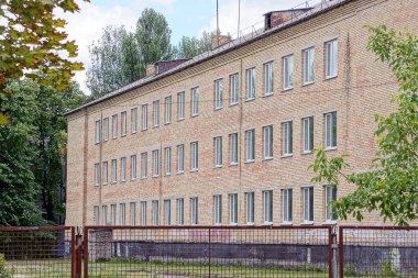 Yeşil bitki örtüsünde, şehir caddesinde bir sıra penceresi olan büyük kahverengi tuğla bir bina duvarı.