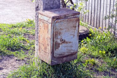 Kahverengi pas içinde gri metal bir kutu ve dışarıda beton bir direğe asılı elektrik kabloları.