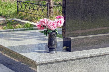 Mezarlıktaki bir mezarın üzerindeki granit bir anıtta kırmızı çiçekli siyah bir mermer vazo.