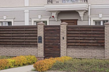 Kapalı kahverengi ahşap kapı. Tuğla ve tahtalardan yapılmış bir duvarın üzerinde. Gri özel bir evin önündeki yeşil çimlerin içinde.