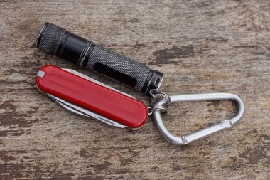Siyah küçük bir el feneri ve gri bir demir karabinanın üzerinde kırmızı bir katlanan bıçak gri bir ahşap masada yatıyor.