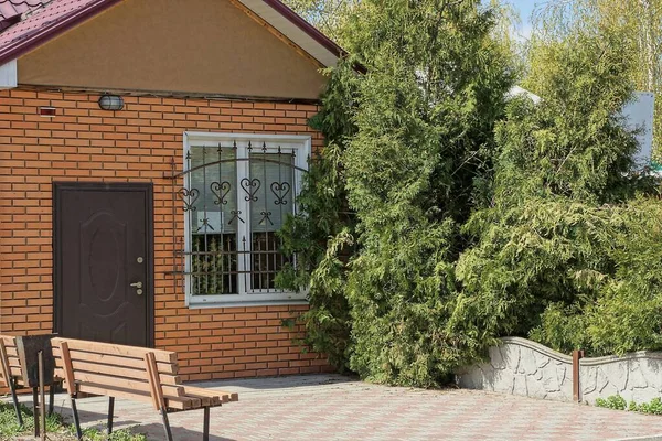 Kahverengi tuğlalı özel bir evin ön cephesinde demir ızgaralı bir pencere ve yeşil kozalaklı ağaçların yanında bir bankı olan gri kaldırımda bir kapı.