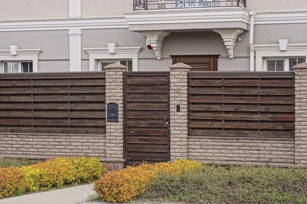 Kapalı kahverengi ahşap kapı. Tuğla ve tahtalardan yapılmış bir duvarın üzerinde. Gri özel bir evin önündeki yeşil çimlerin içinde.