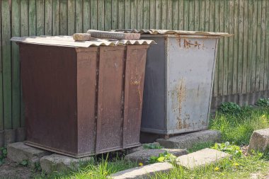 İki demir kahverengi çöp tenekesi dışarıda, yeşil çimlerin arasında tahta bir çitin karşısında duruyor.