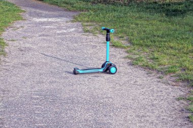Yolun gri asfaltında, parktaki yeşil çimlerin yanında boş mavi bir scooter duruyor.