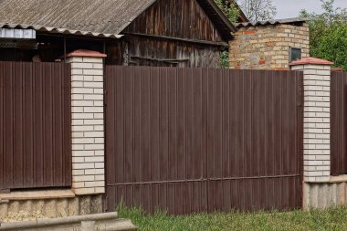 Beyaz tuğlalardan ve yeşil çimenlikteki kırsal bir sokakta demirden yapılmış bir çit duvarındaki büyük kahverengi kapalı metal kapı.