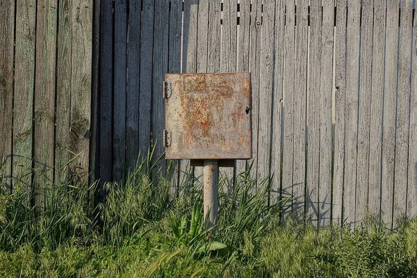 Bir borunun üzerindeki paslı elektrik kabloları için bir kare gri metal kutu sokağın yeşil çimlerinde, tahta bir çitin yanında duruyor.
