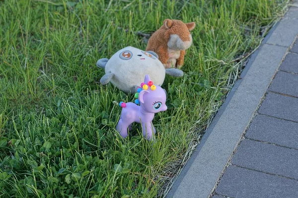a set of three toys with a purple plastic unicorn and teddy bears in the green grass near the gray sidewalk in the park