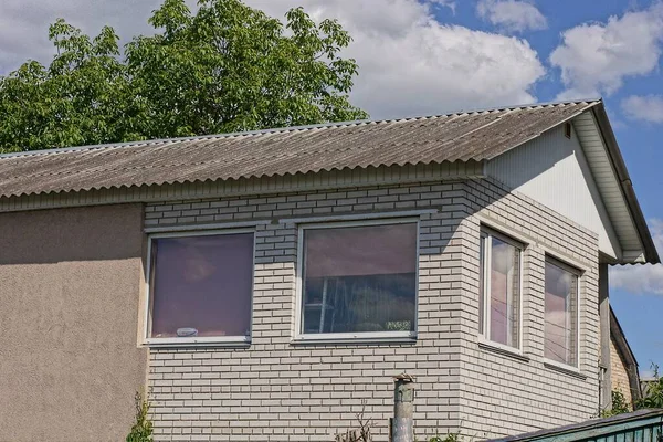 Gökyüzünün ve yeşil ağaçların arka planına karşı gri bir çatı altında pencereleri olan beyaz tuğlalı bir evin tavan arası.