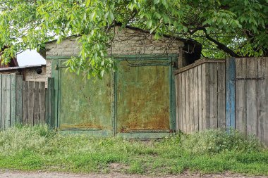 Kırsal beyaz tuğlalı garajın ön cephesinde paslı yeşil metal kapılar ve caddede gri ahşap bir çit var.