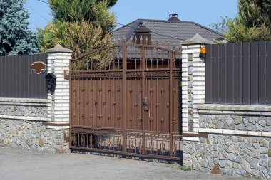 Gri metal ve beyaz tuğlalardan yapılmış bir çitin duvarında dövmeli demir desenli kahverengi, kapalı bir metal kapı var.