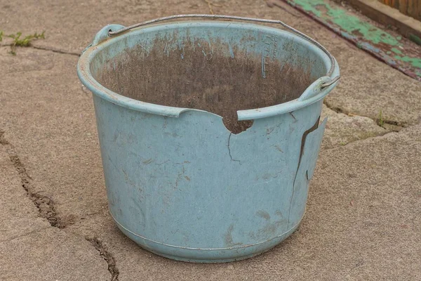 one old blue broken dirty plastic bucket with cracks stands on gray asphalt in the street