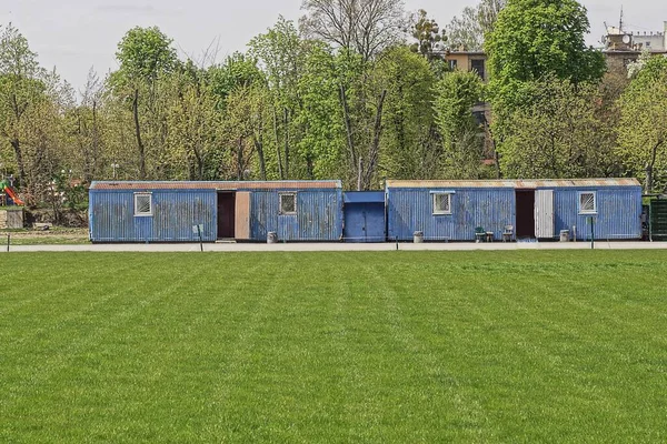 Caddedeki stadyumun yeşil çimlerinin yanında pencereli iki mavi metal römork duruyor.