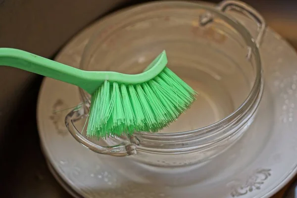 キッチンに緑のプラスチックブラシで汚れた白いプレートとガラスカップをきれいにします — ストック写真