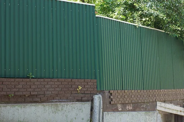 green metal fence wall on a foundation of brown bricks and gray concrete on a rural street