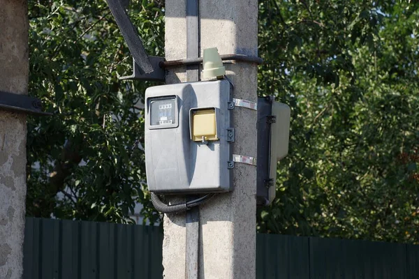 one gray electric meter hangs on a concrete pole in the street against the backdrop of a fence and green vegetation