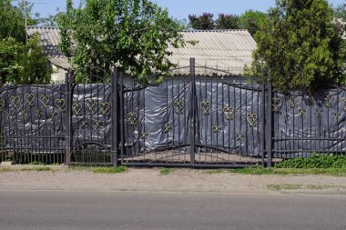 Siyah metal kapı ve demirden yapılmış demir parmaklıklardan yapılmış bir kapı ve caddede gri asfalt bir yolun yanında selofanla kaplı çit duvarın bir kısmı.