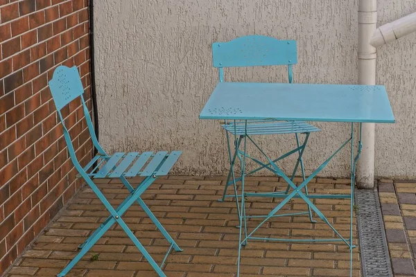 blue metal table and chairs stand on gray brown sidewalk against the wall of the building on the street in a summer cafe