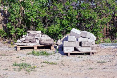 İki ahşap palet, beyaz tuğlalar ve beton bloklar, sokaktaki yeşil bitki örtüsünün arasında.