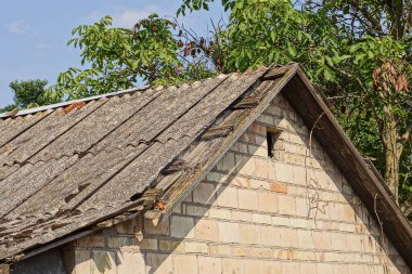 attic of an old brown brick house with a small window and a broken gray slate roof on a rural street against the backdrop of green vegetation and a blue sky
