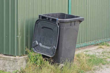 Bir açık siyah plastik çöp tenekesi çimlerin üzerinde yeşil metal bir çit duvarına karşı duruyor.