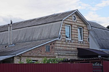 Kahverengi tuğlalı, mavi gökyüzü ve beyaz bulutların arka planına karşı caddede kırmızı metal bir çitin arkasındaki gri tahta çatının altında pencereleri olan kırsal bir ev.