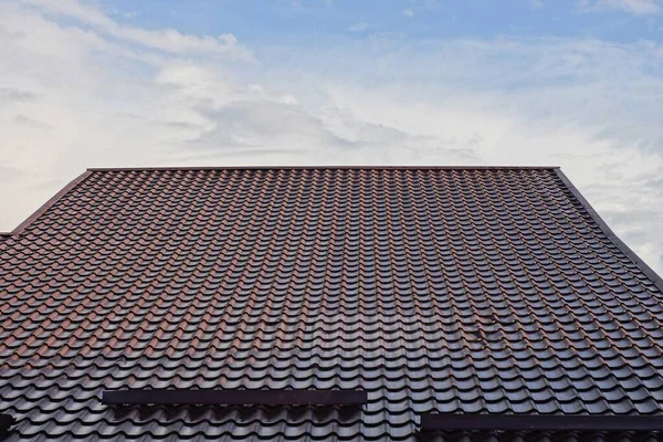 part of the roof of a private house from a brown tiled roof on the street against the background of a blue sky and gray clouds