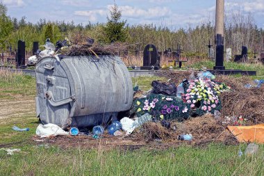 Bir gri metal çöp tenekesi, mezarlığın yanındaki sokakta cenaze çelenkleri olan bir çöp yığınının yanında duruyor.