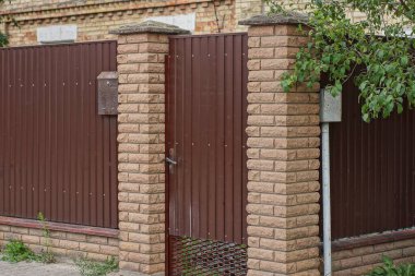 one closed iron brown door and part of a wall fence made of bricks and metal on a rural street