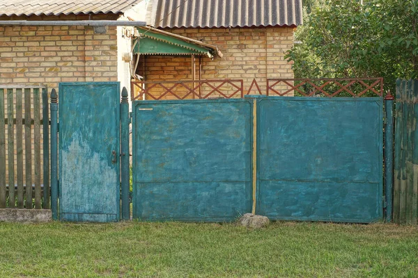 old blue iron gate and a closed metal door on a wooden fence wall in green grass on a rural street