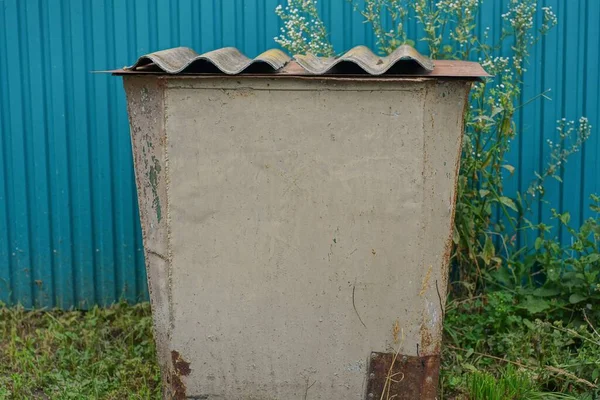 one large gray metal trash can stands in the green grass on the street against the blue wall of the fence