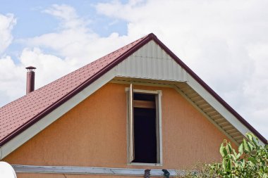 one brown attic of a private house with an open door under a red tiled roof on the street against the background of a blue sky and white clouds