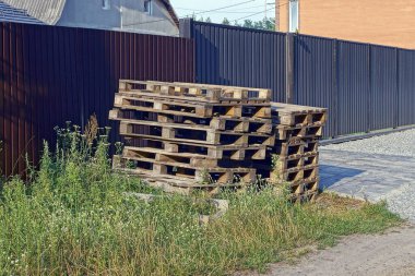 a pile of old brown wooden pallets in green grass on the street near the fence wall