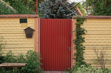 one red closed metal door on a brown concrete fence wall on a rural street in green vegetation