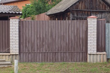 one big brown closed metal gate on a fence wall made of white bricks and iron on a rural street in green grass