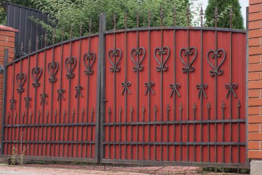 one closed red iron gate with black forged pattern and brown brick wall of the fence on the street