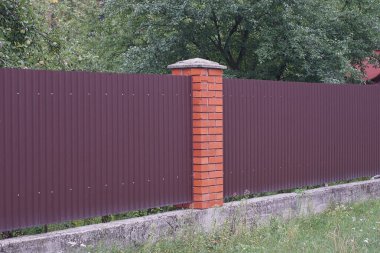 long fence wall made of brown metal and red bricks on a gray concrete foundation in green grass