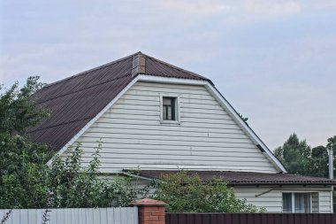 white attic of a rural house with a small window under a gray slate roof on the street against the sky
