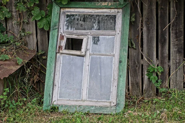 one old wooden window stands on the street in green grass near a gray fence