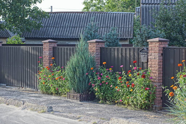 flowers and grass with green coniferous ornamental trees near the brown brick and metal fence wall on a rural street