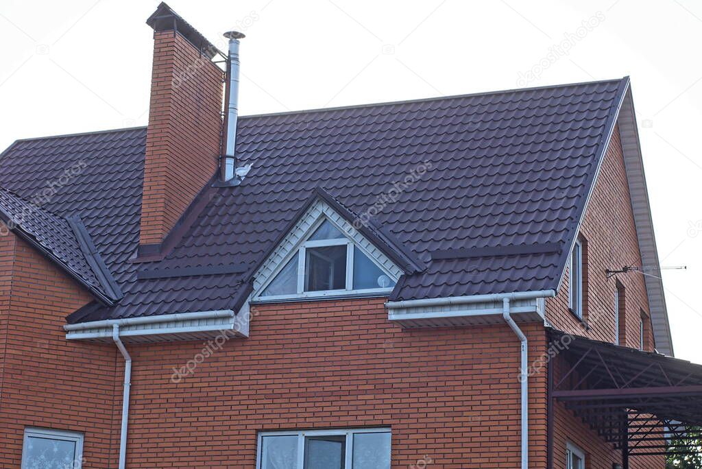 red brick attic with a white windows and one large chimney on a brown tiled roof against a background of gray sky 