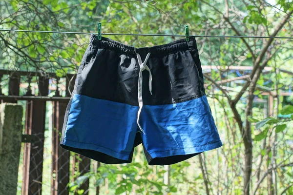 one blue black shorts hanging and drying on a wire with clothespins against the background of green vegetation on the street