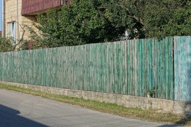 long wooden gray green fence wall on a concrete foundation near an asphalt road in a rural street
