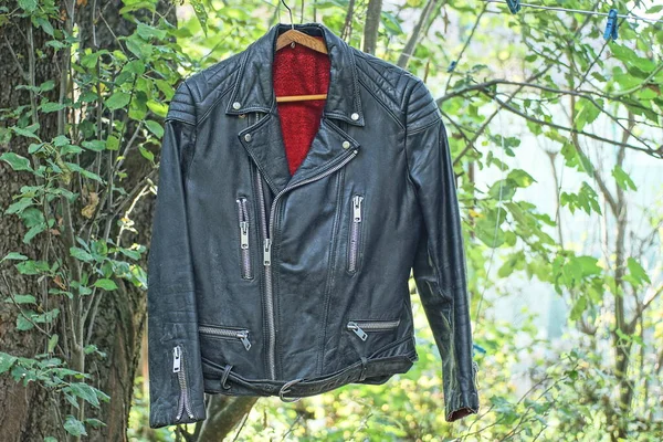 one old black leather jacket with red lining hangs on a wire among the green vegetation on the street