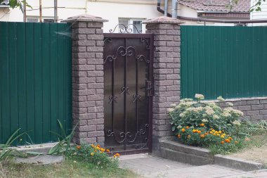one black metal door with wrought iron pattern on the wall of the fence made of brown bricks and green iron in the street